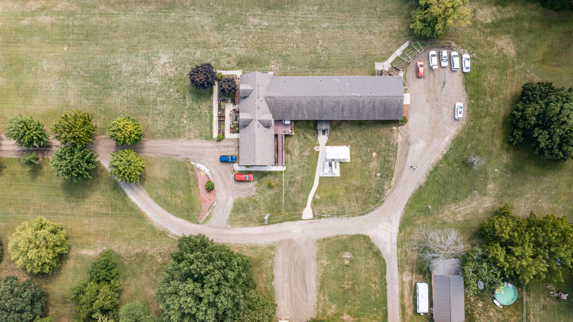 Aerial view of a long building with a curved driveway, parked cars, and surrounding grassy grounds with scattered trees.