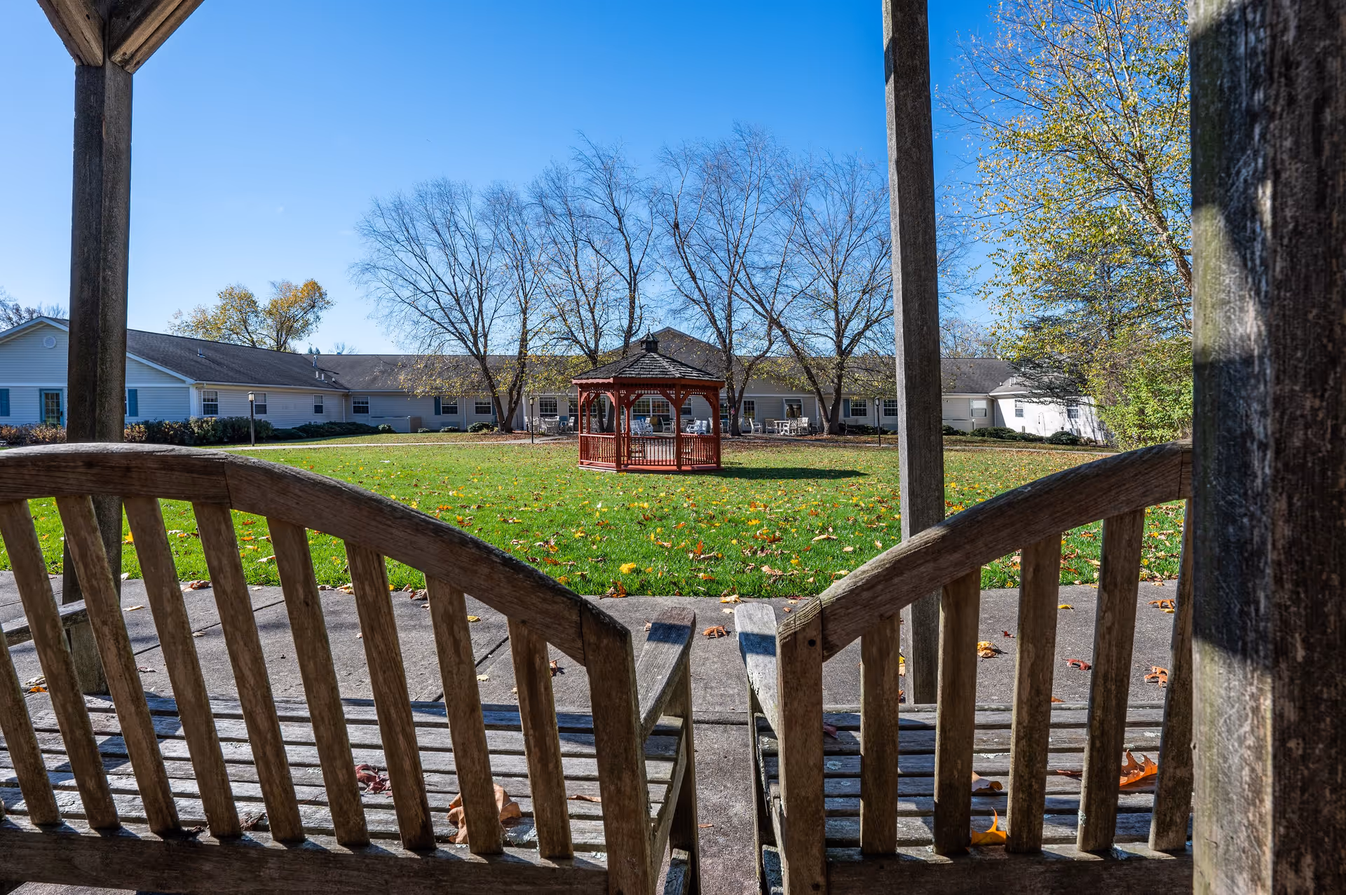 View from a wooden gazebo or covered seating area looking out onto a grassy courtyard with a red gazebo in the center. Surrounding the courtyard are single-story buildings with windows and doors. Trees with sparse leaves are visible, and the sky is clear and blue.