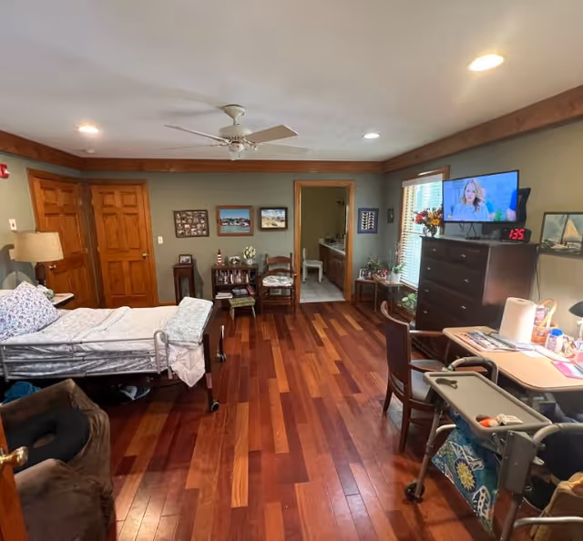 A spacious assisted living room with wooden floors and green walls. The room contains a hospital-style bed with white and floral bedding, a brown recliner chair, a wooden dresser with a TV mounted above it, a clock showing 1:35, and a small table with chairs. There is a ceiling fan and recessed lighting. The room has two wooden doors and an open doorway leading to a bathroom with a white chair visible inside. Various framed pictures and decorations adorn the walls and surfaces.