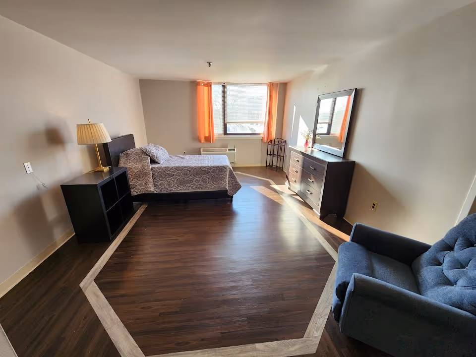 A tidy bedroom in a senior residence featuring a bed, dresser, lamp, and blue chair.