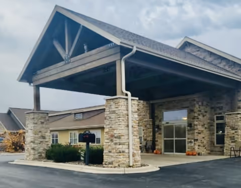 Entrance of a senior care facility with a covered drop-off area supported by stone pillars, a paved driveway, and a building with beige siding and multiple windows in the background under a cloudy sky.