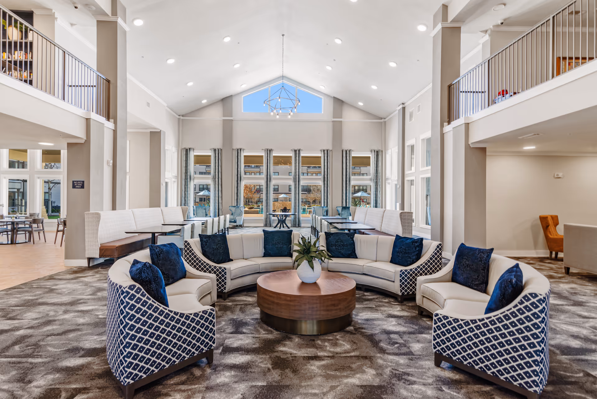 Bright, spacious senior living lounge with curved seating, patterned armchairs, a central round coffee table, and large windows under a vaulted ceiling.