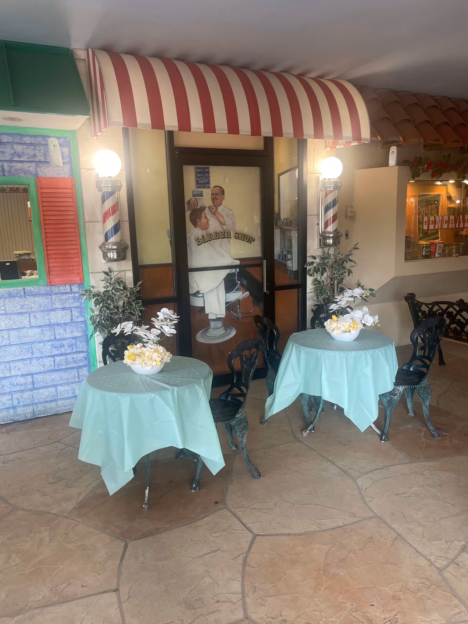 Indoor seating area with two round tables covered with light blue tablecloths, each with a flower arrangement centerpiece. Behind the tables is a door with a painted image of a barber cutting a customer's hair, labeled 'Barber Shop'. The door is flanked by two traditional barber poles with lights on top. To the right, there is a window with a sign that reads 'Dave's General'.