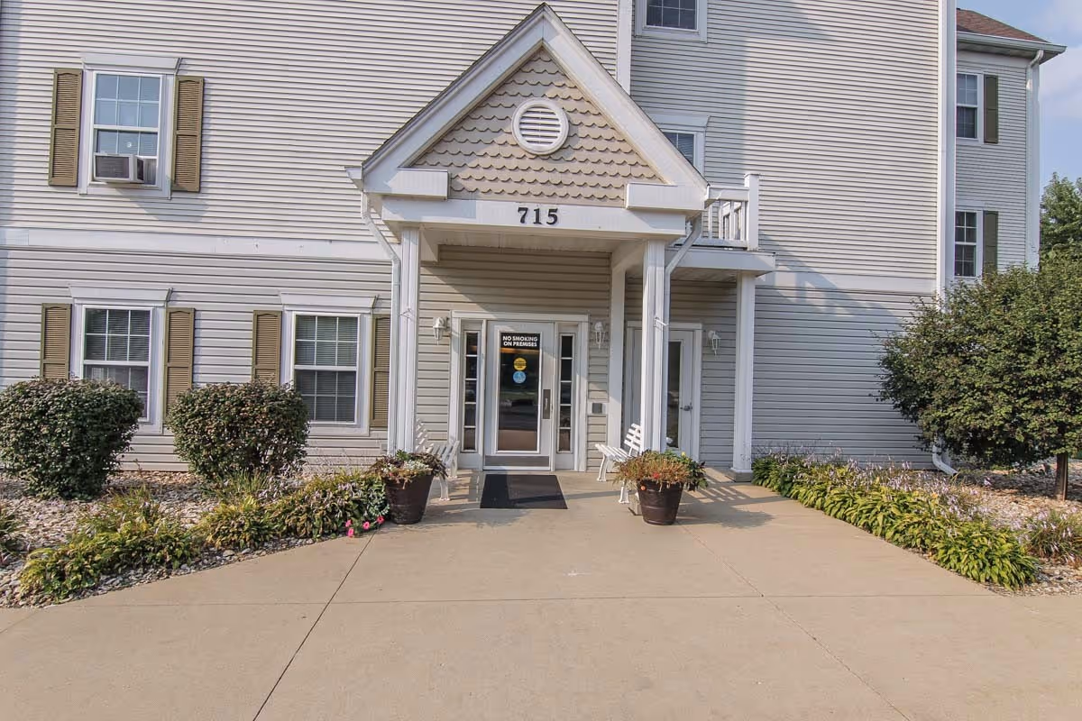 Front entrance of a multi-story beige siding apartment building with the number 715 above the doorway, potted plants, and a concrete walkway.
