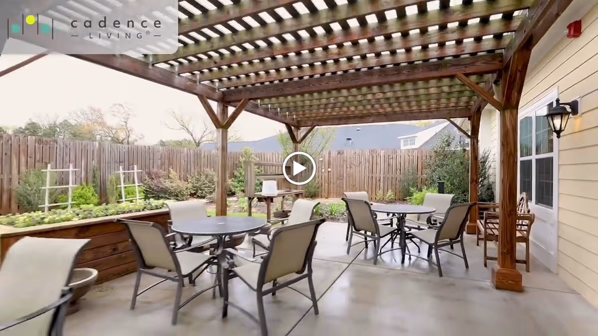 Outdoor patio area with a wooden pergola overhead, several round tables with chairs arranged underneath, a wooden fence surrounding the space, and garden beds with plants along the fence.