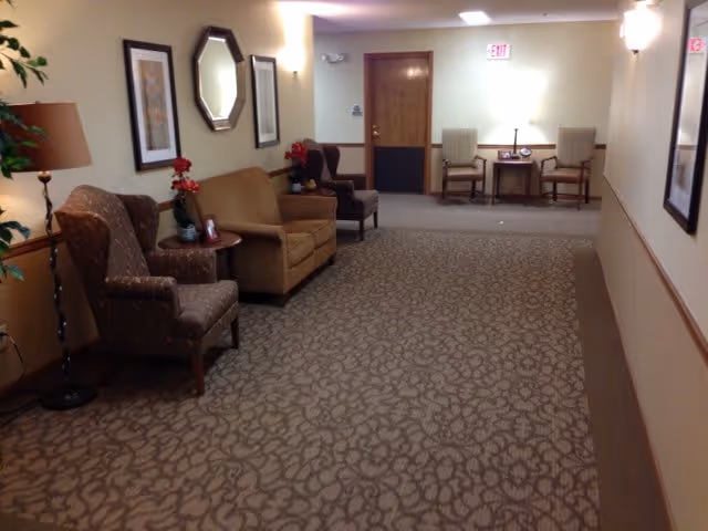 A hallway in a senior living facility with patterned carpet and beige walls. There are several upholstered chairs and small tables with decorative plants and framed artwork on the walls. At the end of the hallway, there is a wooden door and two chairs with a small table between them under a lamp.