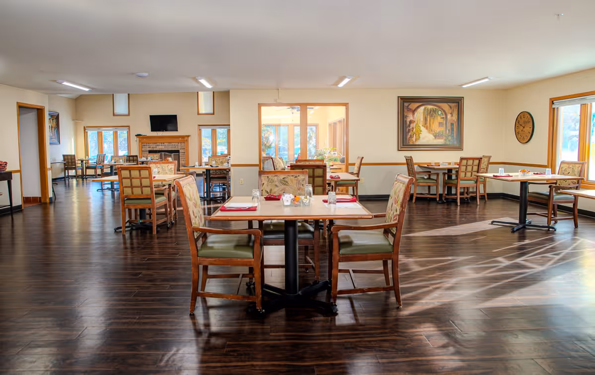 A bright dining room with several wooden tables and chairs arranged neatly. The room features large windows allowing natural light to fill the space, a fireplace with a TV mounted above it, and artwork on the walls. The floor is dark wood, and the tables are set with placemats, napkins, and small decorative items.