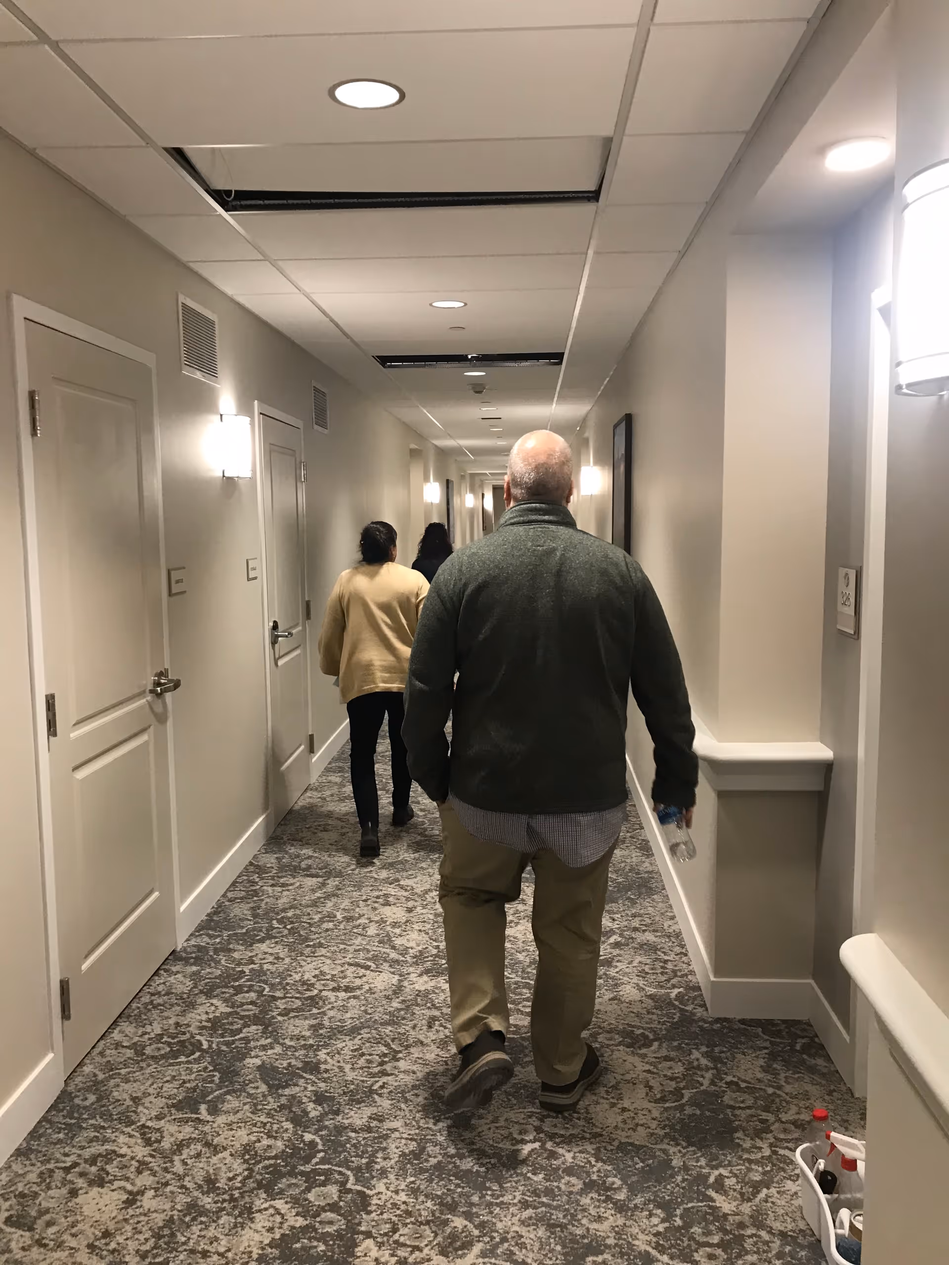 Three people walking down a carpeted interior hallway of a senior living facility.