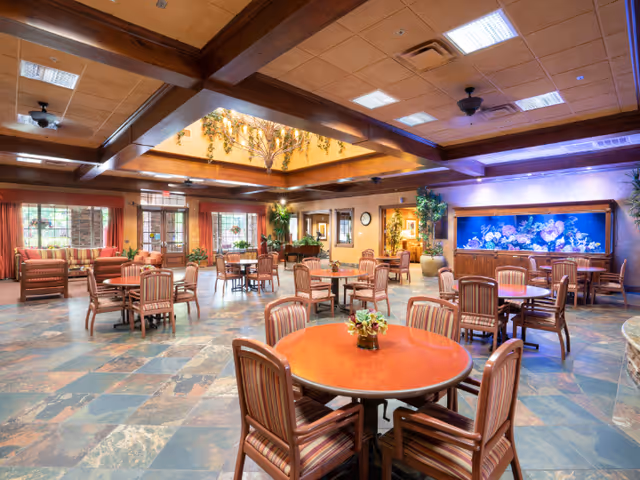 A spacious senior living common area with multiple round tables and striped cushioned chairs arranged throughout the room. The ceiling features wooden beams and a recessed area with decorative plants. Large windows and glass doors allow natural light to enter. There is a large aquarium built into one wall and various plants and seating areas around the room.