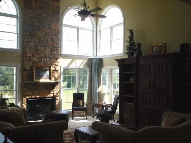 Sunlit living room with stone fireplace, tall arched windows, and dark wooden furniture.