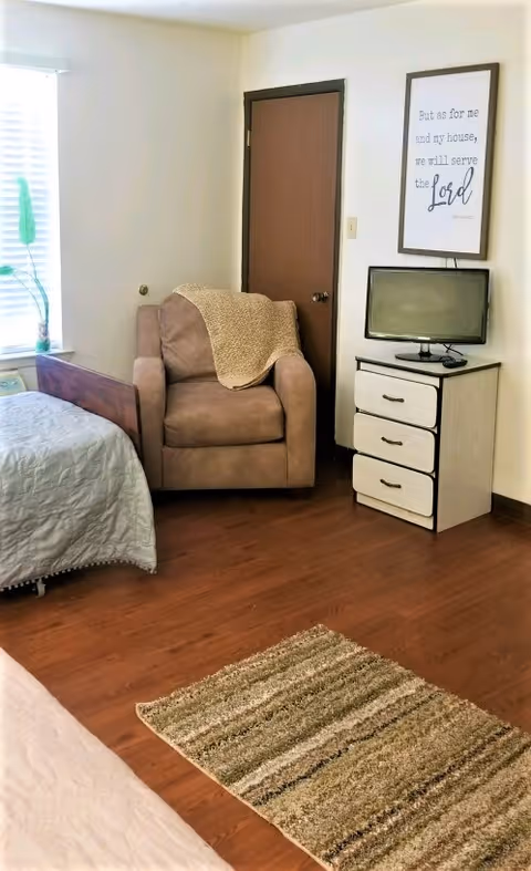 A cozy room with a brown armchair draped with a beige knitted blanket, a small dresser with three drawers holding a flat-screen TV, a framed inspirational quote on the wall, a bed with a light gray quilt, a window with blinds and a green plant on the windowsill, and a striped area rug on the wooden floor.