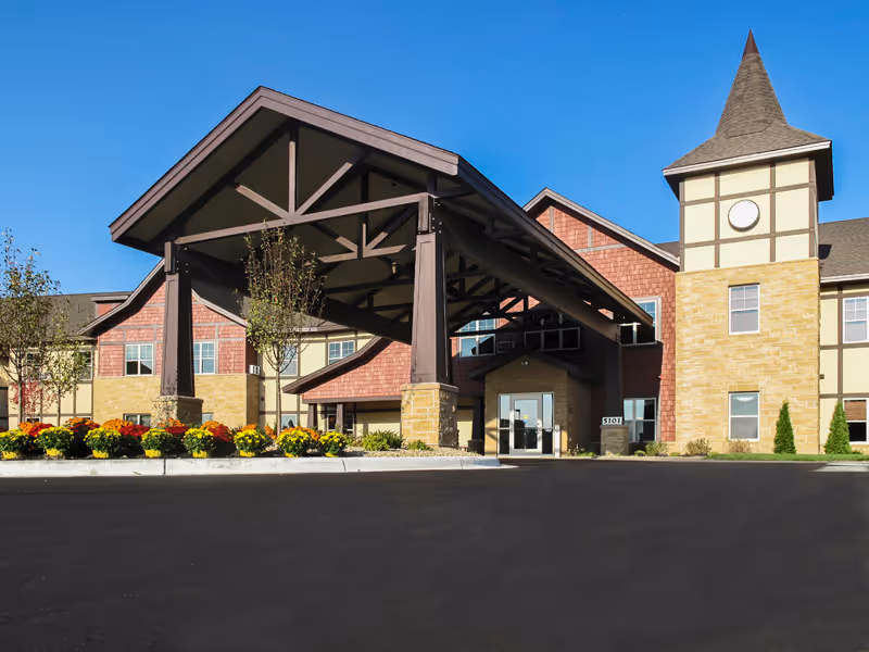 Front exterior view of Guardian Angels Engel Haus Senior Living facility in Albertville, showing the entrance with a large covered driveway, landscaped flower beds, and a tower-like structure on the right side of the building under a clear blue sky.