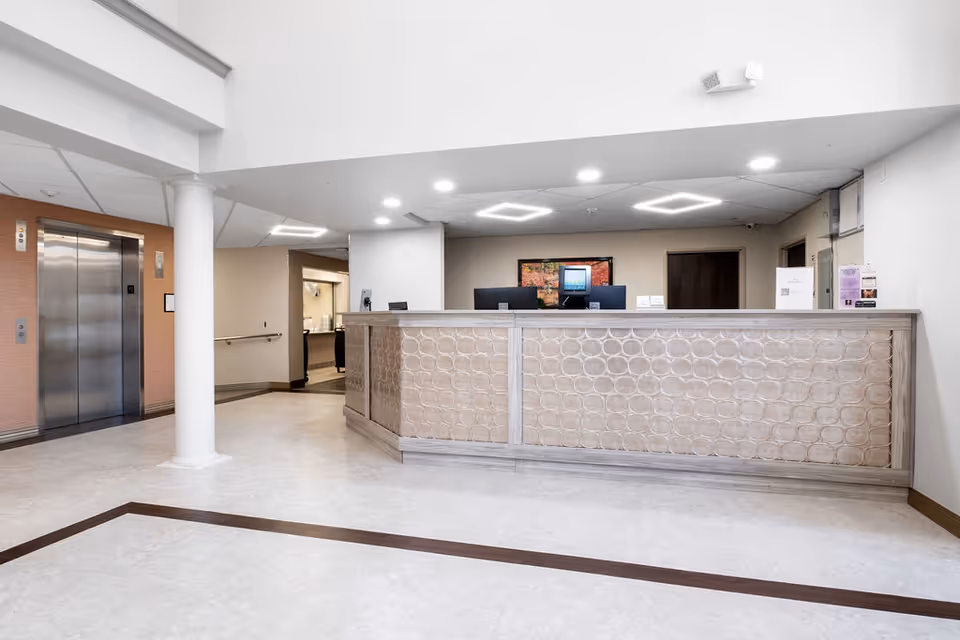 Reception area of Mountain View Senior Living featuring a modern front desk with a textured front panel, two computer monitors, and a framed picture on the wall behind. To the left, there is a stainless steel elevator door and a white column. The floor is light-colored with a dark border design, and the ceiling has recessed and geometric light fixtures.