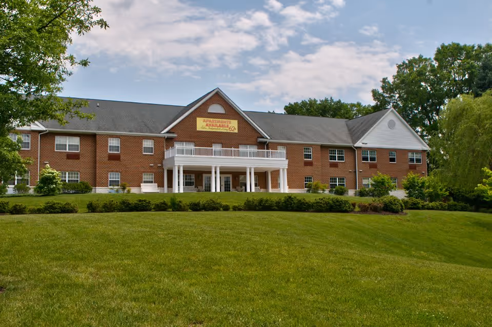 Exterior view of a two-story brick building with white columns and a balcony, surrounded by green grass and trees under a partly cloudy sky. A yellow sign on the building reads 'Apartments Available 62+'.