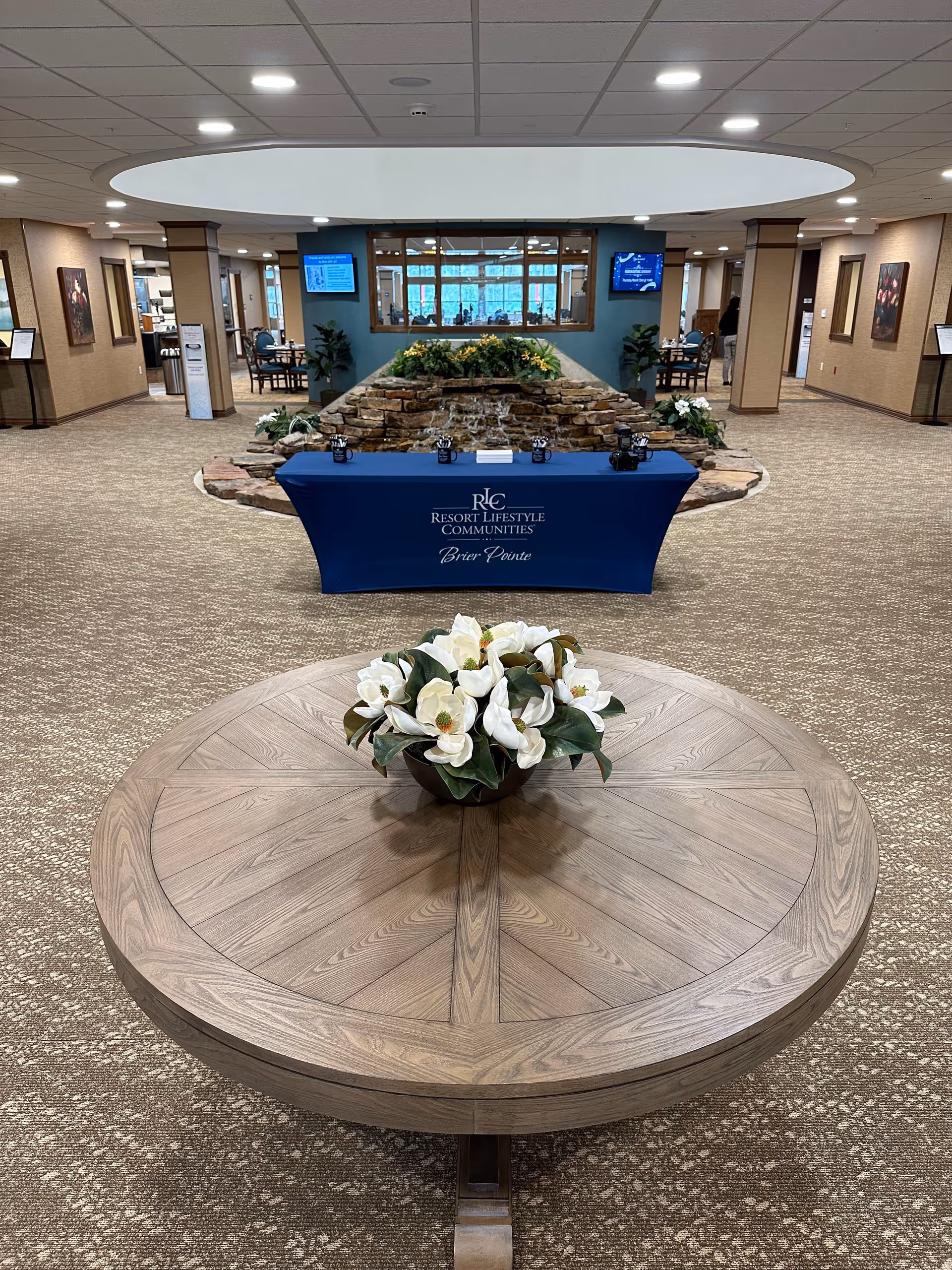 Interior view of a spacious common area in a retirement community featuring a round wooden table with a floral centerpiece in the foreground. Behind the table is a blue table covered with a cloth displaying the Resort Lifestyle Communities logo and the name Brier Pointe. A stone water feature with plants is situated behind the table. The area is well-lit with ceiling lights and has carpeted floors and beige walls with framed artwork. Two digital screens are mounted on the walls in the background.