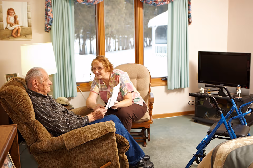 An elderly man sitting in a brown recliner chair and a woman sitting on a cushioned wooden chair in a cozy living room. The woman is holding papers and smiling at the man. Behind them is a large window with light blue curtains showing a snowy outdoor scene. A blue walker is positioned near a television on a stand to the right.