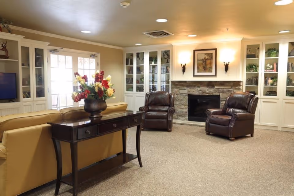 A cozy living room with a stone fireplace centered on the far wall, flanked by built-in white cabinets with glass doors displaying books and decorative items. In front of the fireplace are two dark brown leather armchairs. A tan leather sofa faces away from the fireplace, with a dark wooden console table behind it holding a vase of colorful flowers. The room is softly lit with wall sconces and recessed ceiling lights.
