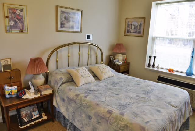 A cozy bedroom with a brass bed frame, a bed covered with a floral patterned comforter and two decorative pillows. There are two wooden nightstands on either side of the bed, each with a lamp and various personal items. The room has three framed pictures on the walls and a window with blinds letting in natural light. A digital clock and framed photographs are visible on the nightstands.