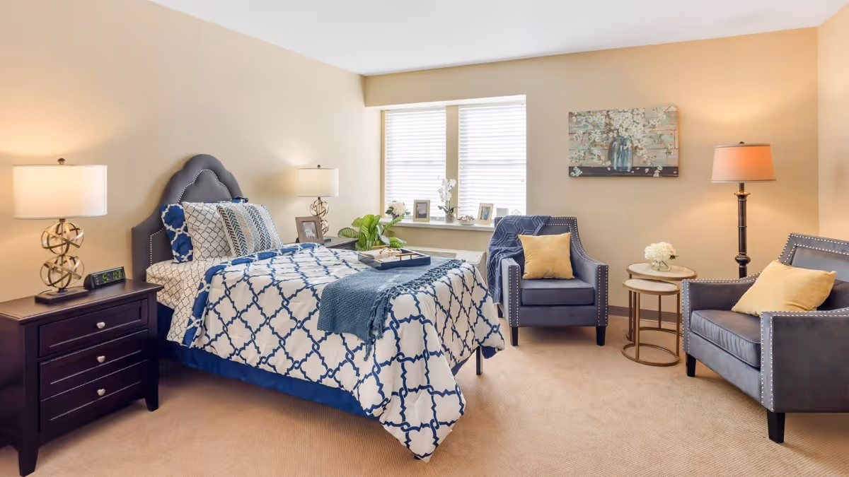 Bright furnished bedroom with a patterned bed, two upholstered chairs, side tables, lamps, and a window.
