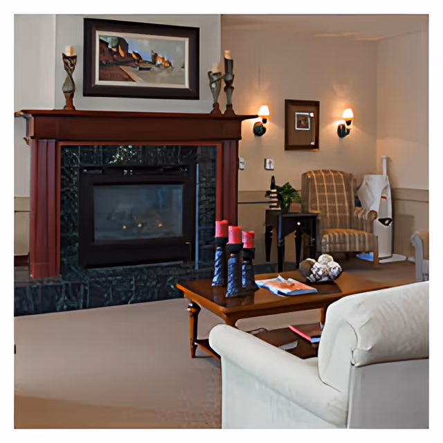 A cozy living room area featuring a fireplace with a dark green marble surround and a wooden mantel adorned with candle holders. Above the mantel hangs a framed landscape painting. In front of the fireplace is a wooden coffee table with decorative candles and a magazine. To the right, there is a striped armchair next to a small side table with a plant and a framed picture on the wall. The room is softly lit by wall sconces.