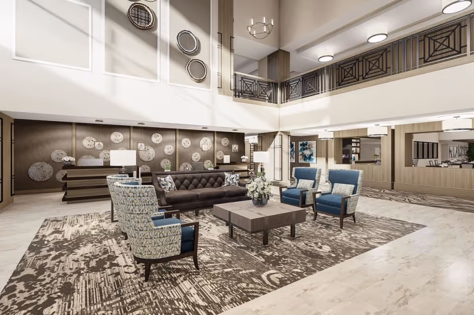 A spacious and well-lit senior living facility lobby with modern decor. The seating area includes a dark tufted sofa, two patterned armchairs, and two blue armchairs arranged around a square coffee table with a floral centerpiece. The floor features a large patterned rug, and the walls are decorated with circular art pieces. The lobby has a high ceiling with a balcony railing above and multiple ceiling lights. A reception desk is visible in the background.
