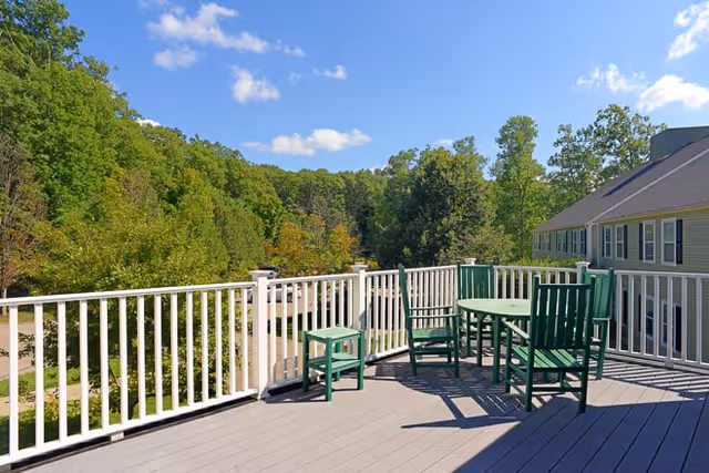 Outdoor deck area with green wooden chairs and a round table, surrounded by white railing. Trees and a building are visible in the background under a blue sky with a few clouds.