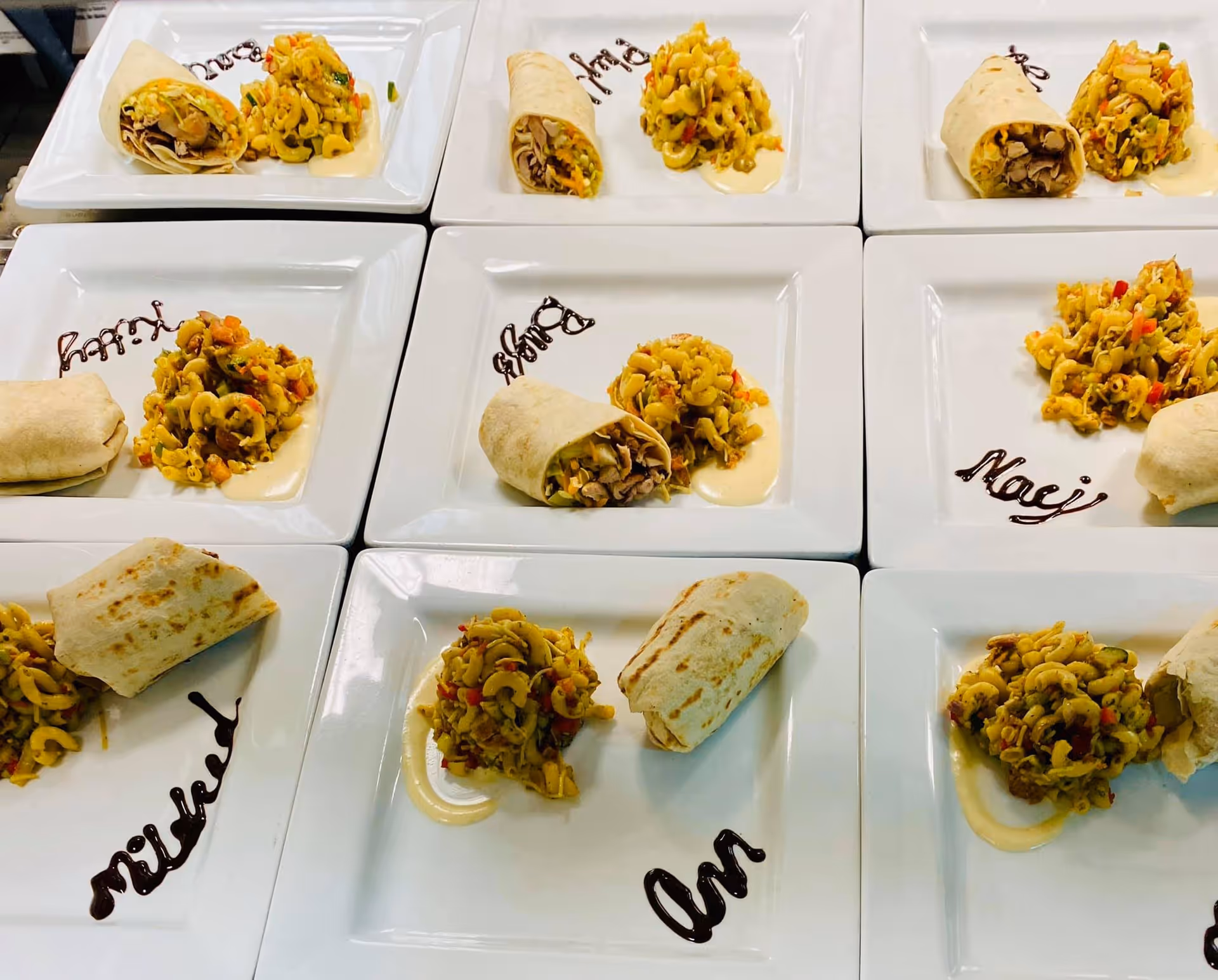 Nine white square plates arranged in a grid, each with a serving of pasta salad and a wrap. Each plate has a name written in chocolate sauce on the side.