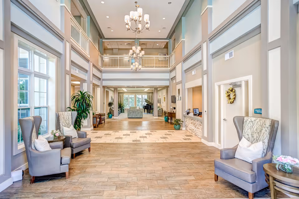 Bright two-story senior living lobby with armchairs, chandeliers, a reception desk and a piano in the distance.