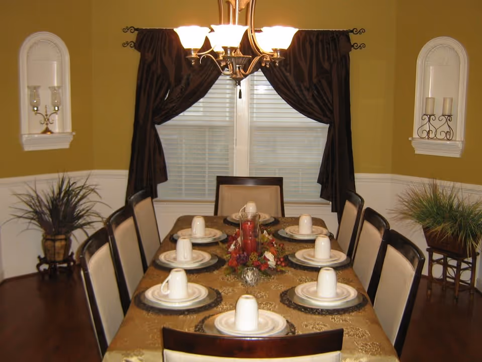 A formal dining room with a rectangular table set for eight people. Each place setting includes a plate, a smaller plate, and an upside-down white cup. The table is covered with a gold-colored tablecloth and has a centerpiece with red candles and decorative flowers. The room has mustard yellow walls with white wainscoting, two windows with dark brown curtains, and a chandelier with five lights hanging above the table. There are decorative wall niches with candle holders and two potted plants on either side of the room.