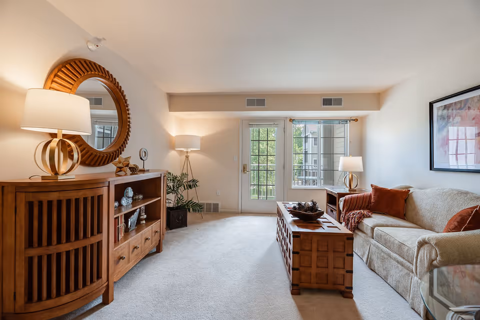 Well-lit living room with a beige couch, wooden coffee table and sideboard with lamps and a round mirror, plus glass doors and windows leading to a balcony.