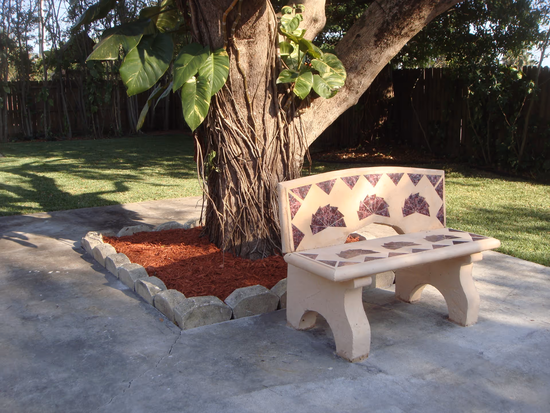 Decorative mosaic stone bench beside a large tree with a mulched planting bed on a concrete patio and lawn.