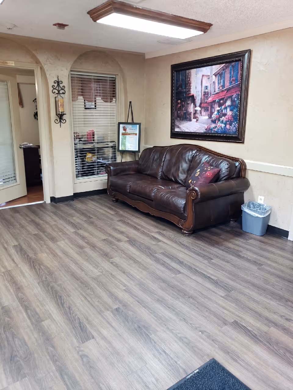 A waiting area with a dark brown leather couch adorned with a decorative pillow. Above the couch hangs a framed painting of a quaint street scene with flowers. The room has wood-look flooring, beige textured walls, a small trash bin next to the couch, and a window with blinds. There is a doorway leading to another room and a decorative wall sconce with a candle on the left wall.