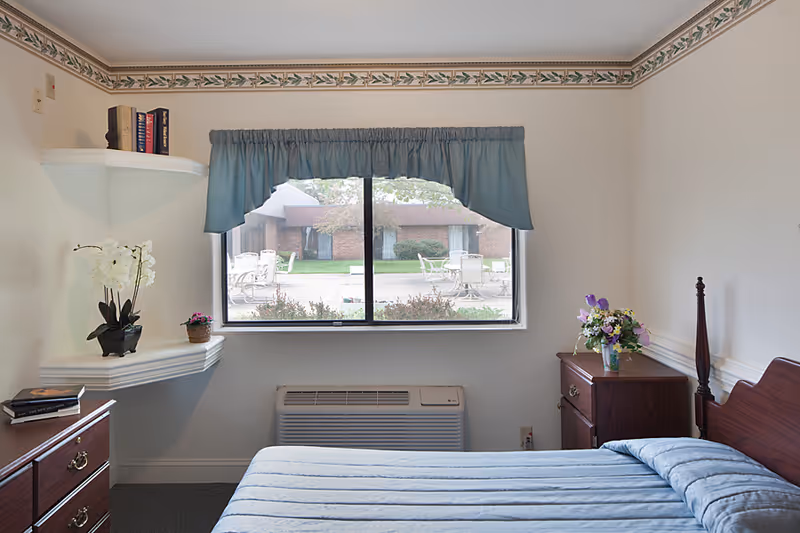 A senior living community bedroom with a bed covered in a blue quilt, a wooden nightstand with a vase of flowers, a wooden dresser with books, a window with a blue valance showing an outdoor patio area with tables and chairs, and white walls with a decorative border near the ceiling.