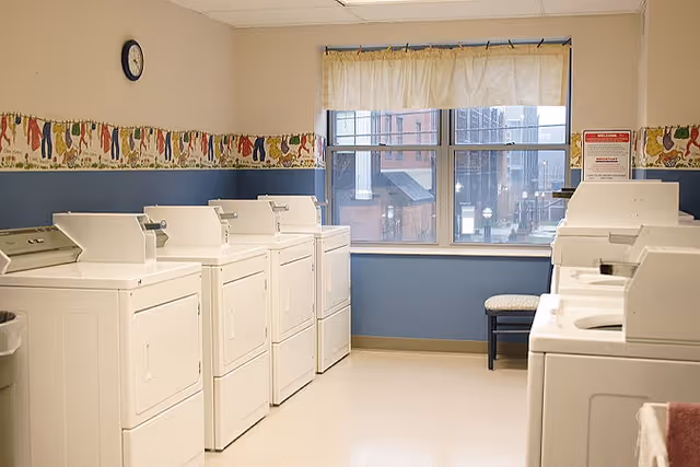 Laundry room with multiple washers and dryers lined along the walls beneath a window and a small bench.