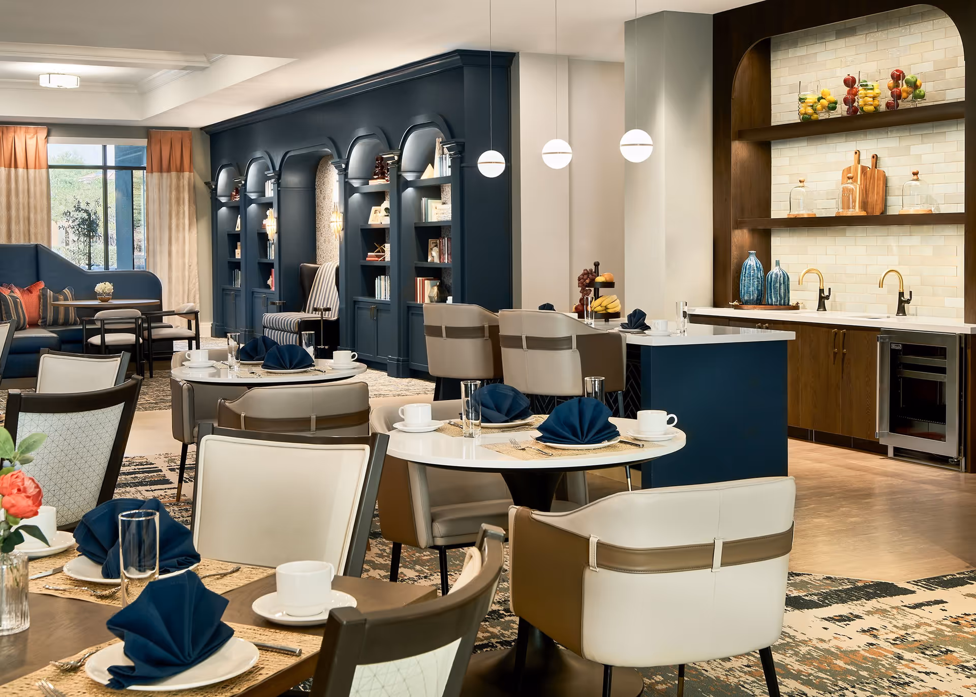Open dining and lounge area in a senior living facility with set tables, upholstered chairs, a small kitchen/bar, and built-in book-lined shelving.