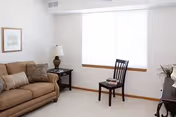 A bright living room with a beige sofa adorned with striped and solid cushions, a dark wooden side table with a lamp, a wooden chair with a book on the seat, a large window with white blinds, and a framed picture on the wall.