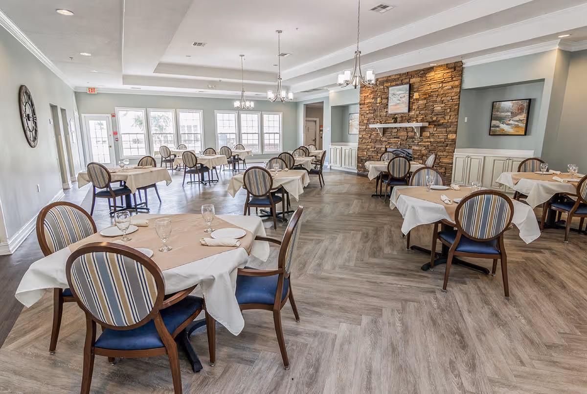 A spacious dining room with multiple round tables covered with white and beige tablecloths, each set with plates, napkins, and glasses. The room features wooden chairs with striped upholstery, large windows letting in natural light, a stone accent wall with a fireplace, and framed artwork on the walls. The floor has a light wood pattern, and several chandeliers hang from the ceiling.