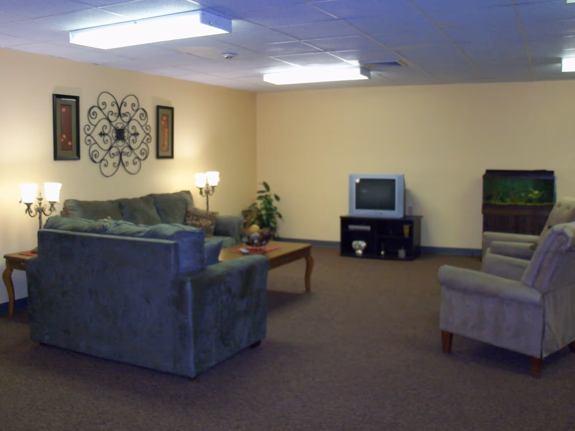 A living room with beige walls and carpeted floor featuring a green sofa set, two armchairs, a wooden coffee table with a decorative bowl, two table lamps, a small TV on a black stand, a fish tank, and wall decorations including framed pictures and a metal wall art piece.