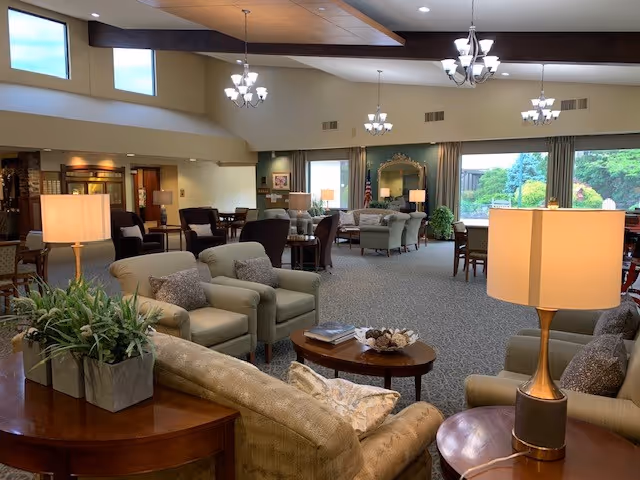 A spacious and well-lit living room area in a senior living facility with comfortable armchairs and sofas arranged around coffee tables. The room features large windows with views of greenery outside, multiple chandeliers hanging from the ceiling, and decorative plants. There are also lamps on side tables providing warm lighting.