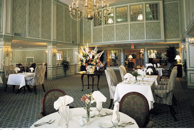 Elegant dining room with multiple tables covered in white tablecloths, each set with folded napkins, glassware, and silverware. The room features patterned green wallpaper, carpeted floors, a large floral centerpiece on a round table, and a chandelier hanging from the ceiling.