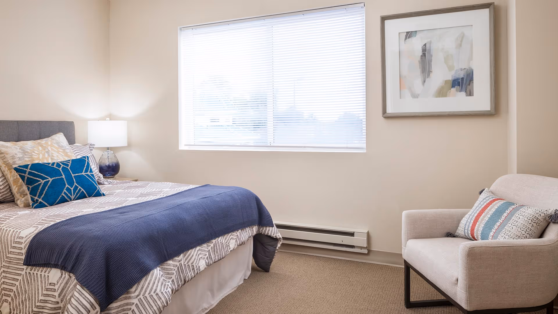 A bright bedroom with a large window covered by white blinds. The room features a bed with patterned bedding, a blue throw blanket, and decorative pillows. Next to the bed is a nightstand with a lamp. There is a beige armchair with a striped pillow and a framed abstract painting on the wall.