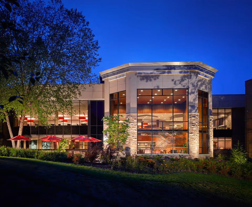 Dusk exterior view of a modern senior living building with large illuminated glass windows, visible interior stairwell, red patio umbrellas, and landscaped grounds.