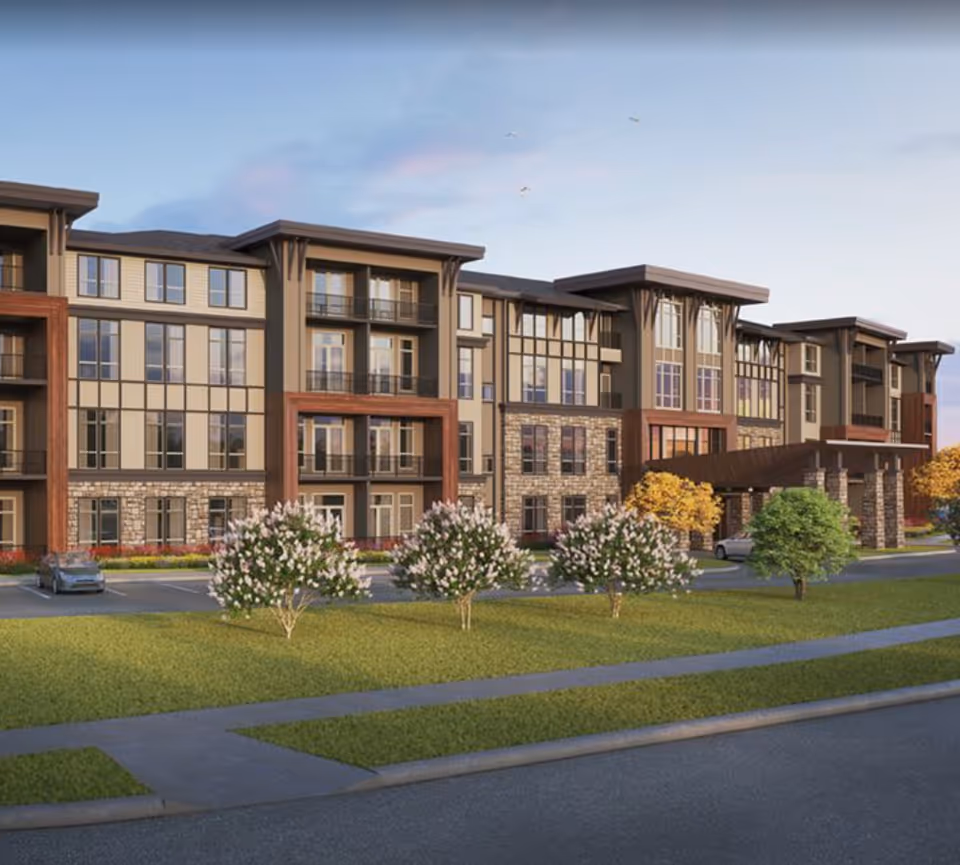 Exterior view of a modern multi-story senior living facility with large windows, balconies, stone and wood accents, surrounded by green lawns, flowering bushes, and trees under a clear sky.