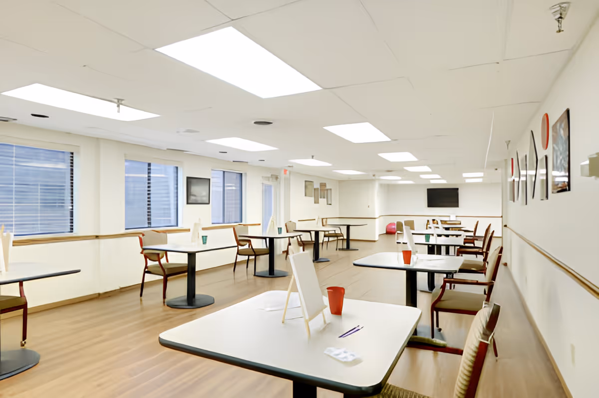 Bright, spacious dining/activity room with multiple tables and chairs, large windows, and a wall-mounted TV.
