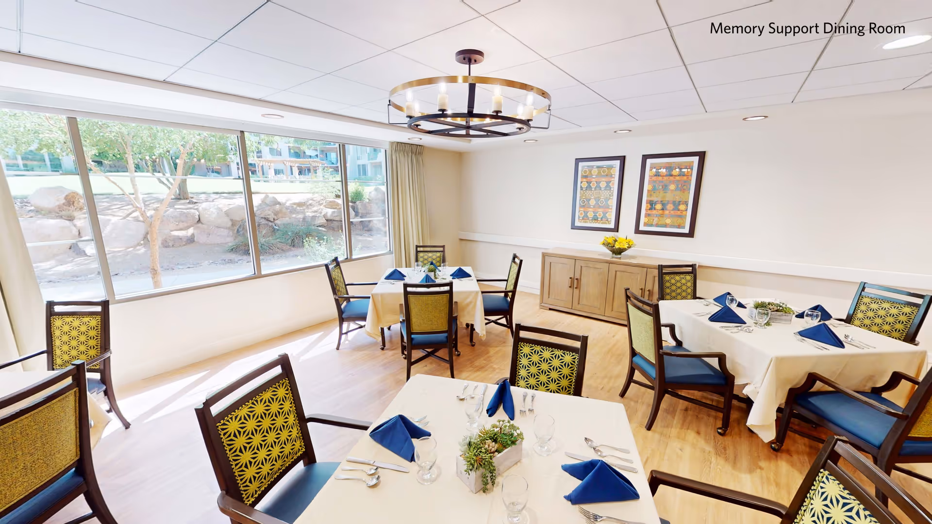 Bright dining room with several round tables set with blue napkins and chairs, large windows overlooking landscaping, and wall artwork.