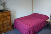 Small bedroom with a double bed covered in a red patterned bedspread, a wooden dresser, and a bedside table.
