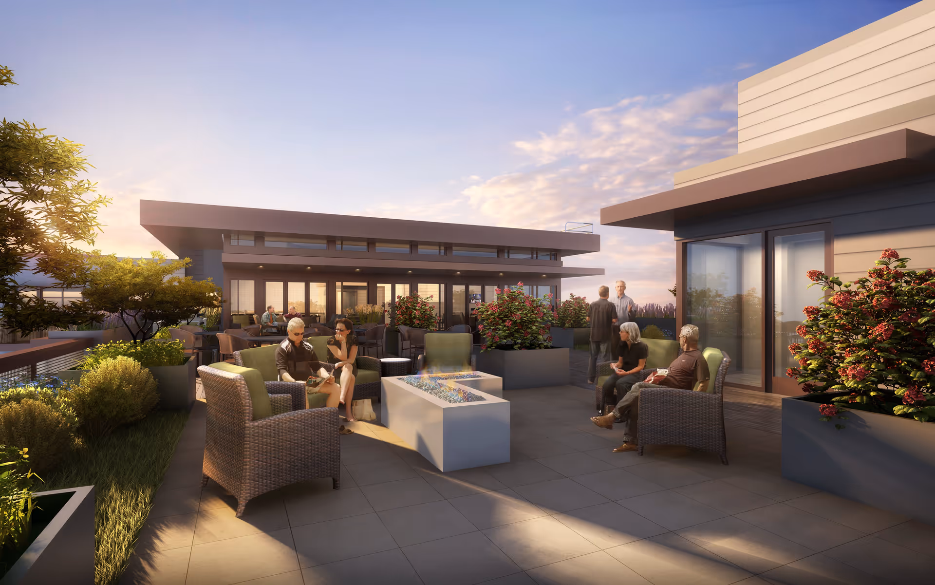 Outdoor patio area at a senior living facility with several people sitting and conversing on wicker chairs around a modern rectangular fire pit. The patio is decorated with large planters filled with green shrubs and flowering plants. The building has large windows and a modern architectural design, with a clear sky and soft evening light.