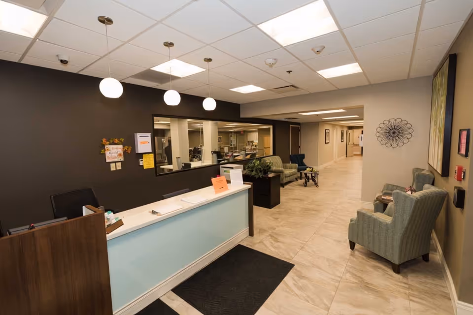 Reception area of Keystone Ridge Post Acute Nursing and Rehabilitation featuring a front desk with three hanging pendant lights above, a dark accent wall behind the desk, and a large window looking into another room. The area has tiled flooring, several upholstered chairs arranged along the hallway, decorative wall art, and a long corridor leading further into the facility.