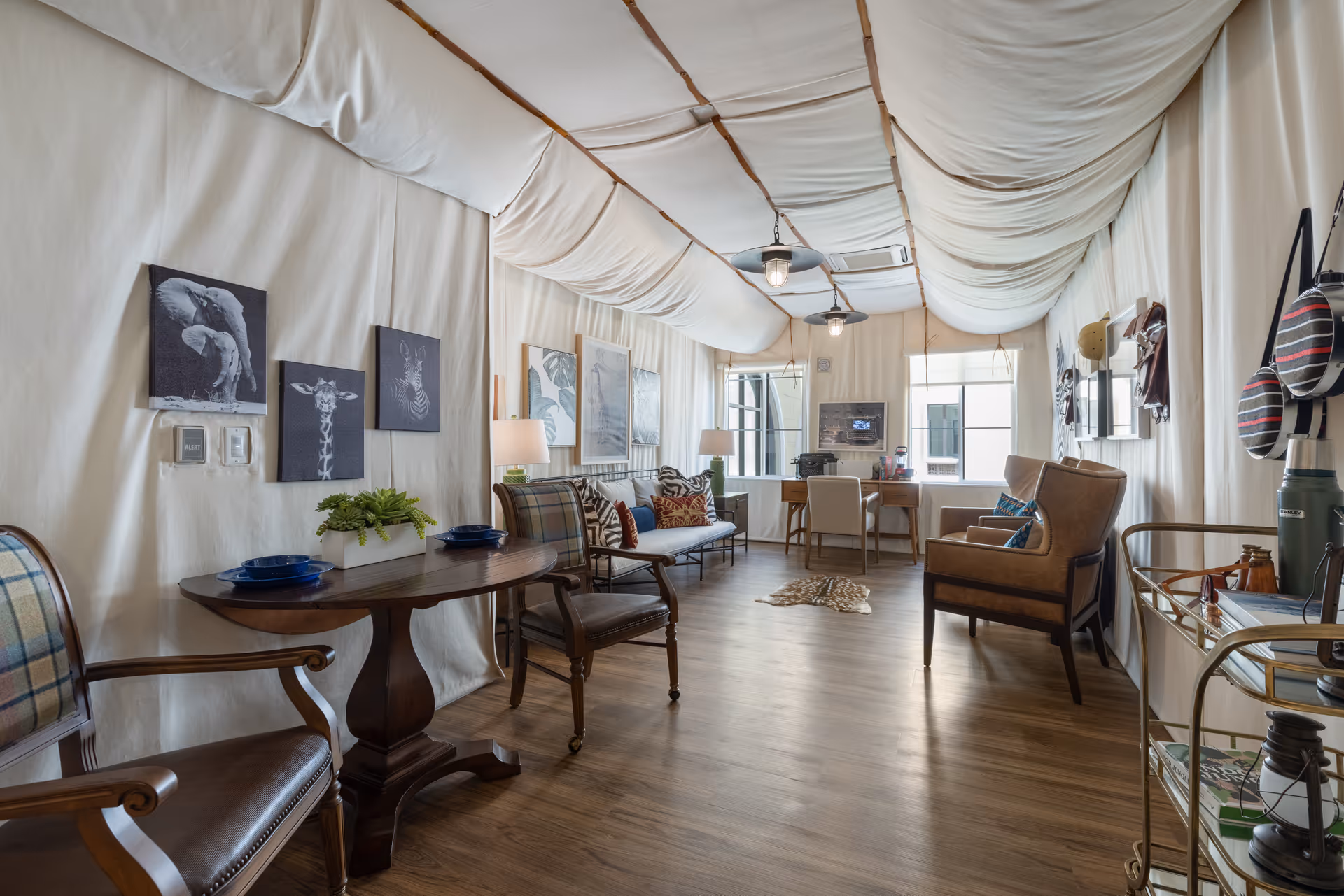 Bright, well-decorated communal living room with a draped fabric ceiling, multiple seating areas, wooden floors, artwork on the walls, and a desk by the windows.
