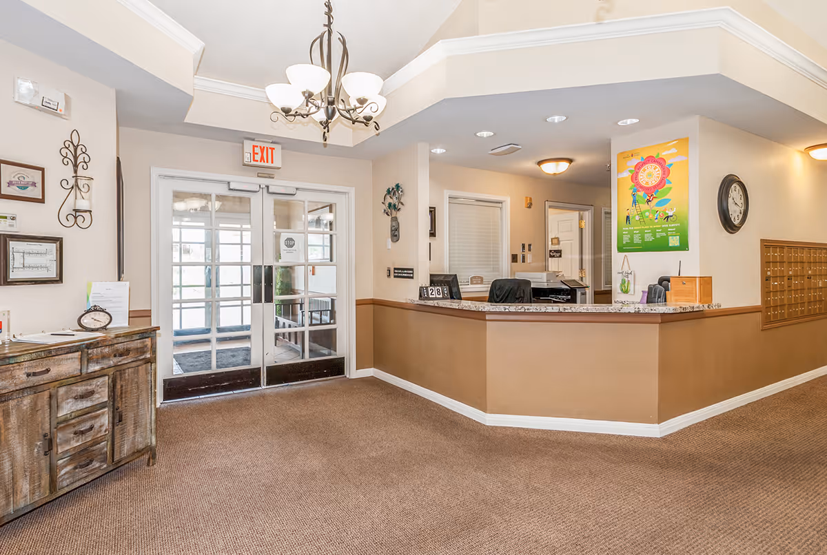 Reception area of Avista Senior Living Apache Junction featuring a front desk with office supplies, a wall clock, mailboxes, a chandelier, and double glass doors with an exit sign above them.